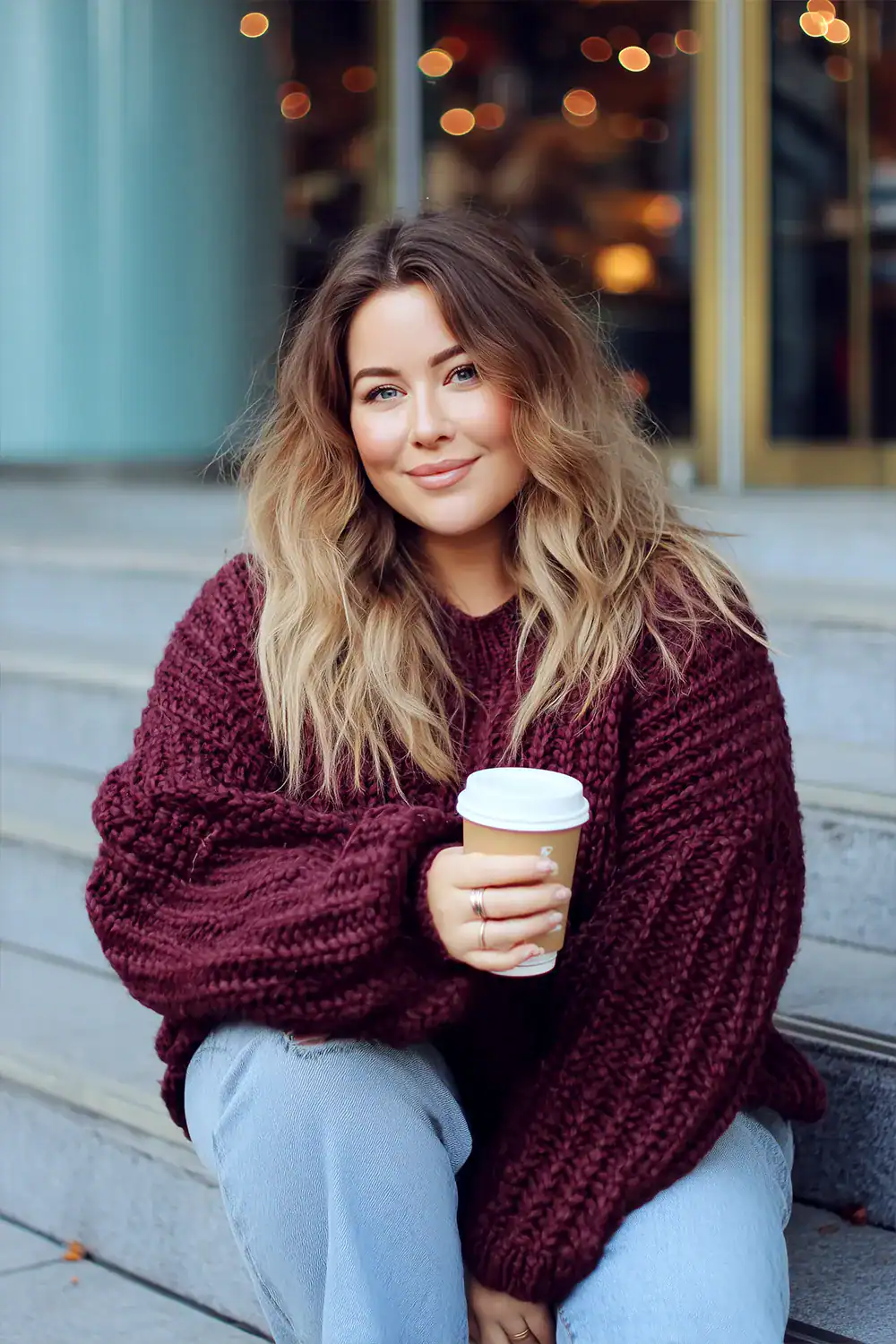 Lächelnde Frau in oversized Strickpullover und Jeans mit Coffee-to-go auf einer Treppe im herbstlichen Stadtambiente