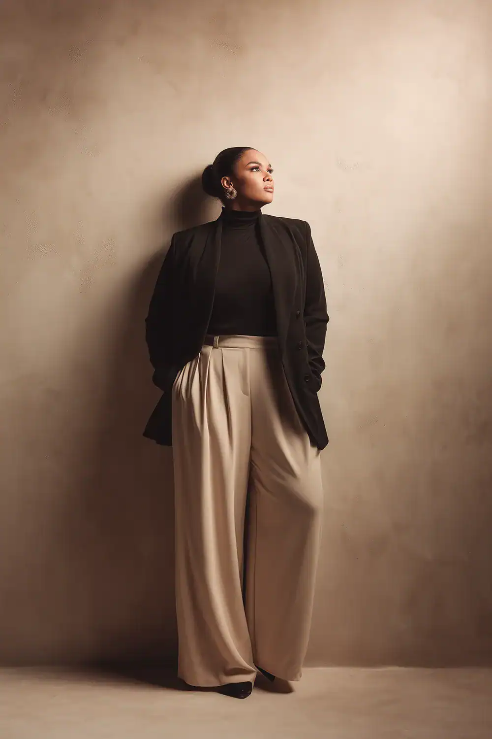 Elegante Plus Size Frau im schwarzen Blazer, Rollkragen und beiger Wide-Leg-Hose vor neutralem Hintergrund