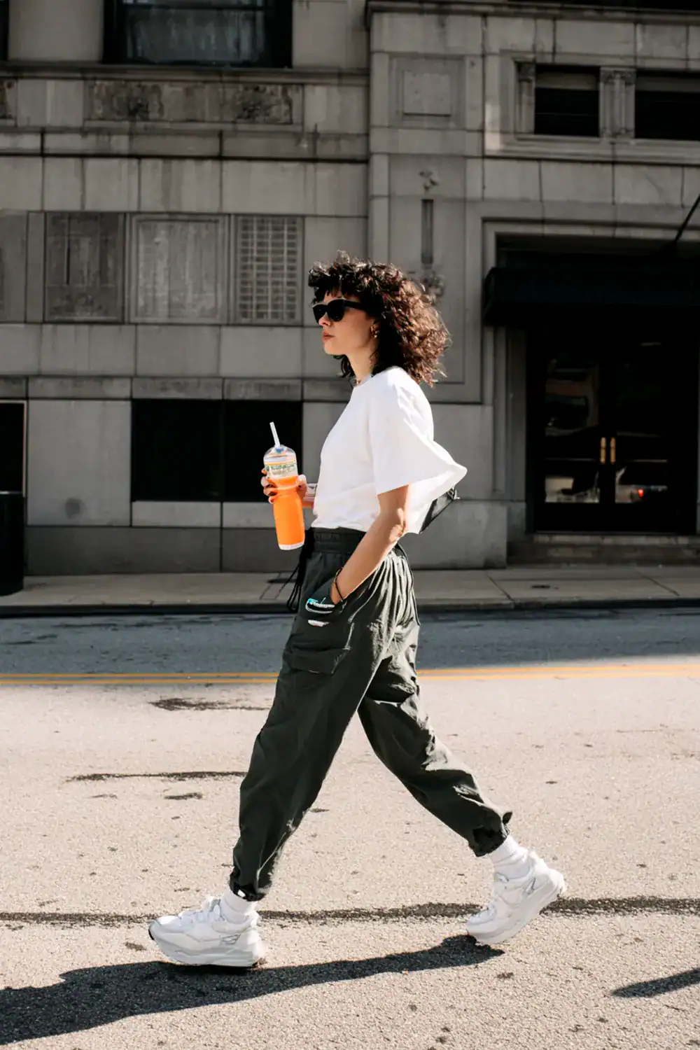 Frau im Streetstyle-Look mit weißen Sneakern, Cargo-Hose, weißem T-Shirt und orangefarbenem Getränk, läuft auf der Straße unter Sonnenlicht.