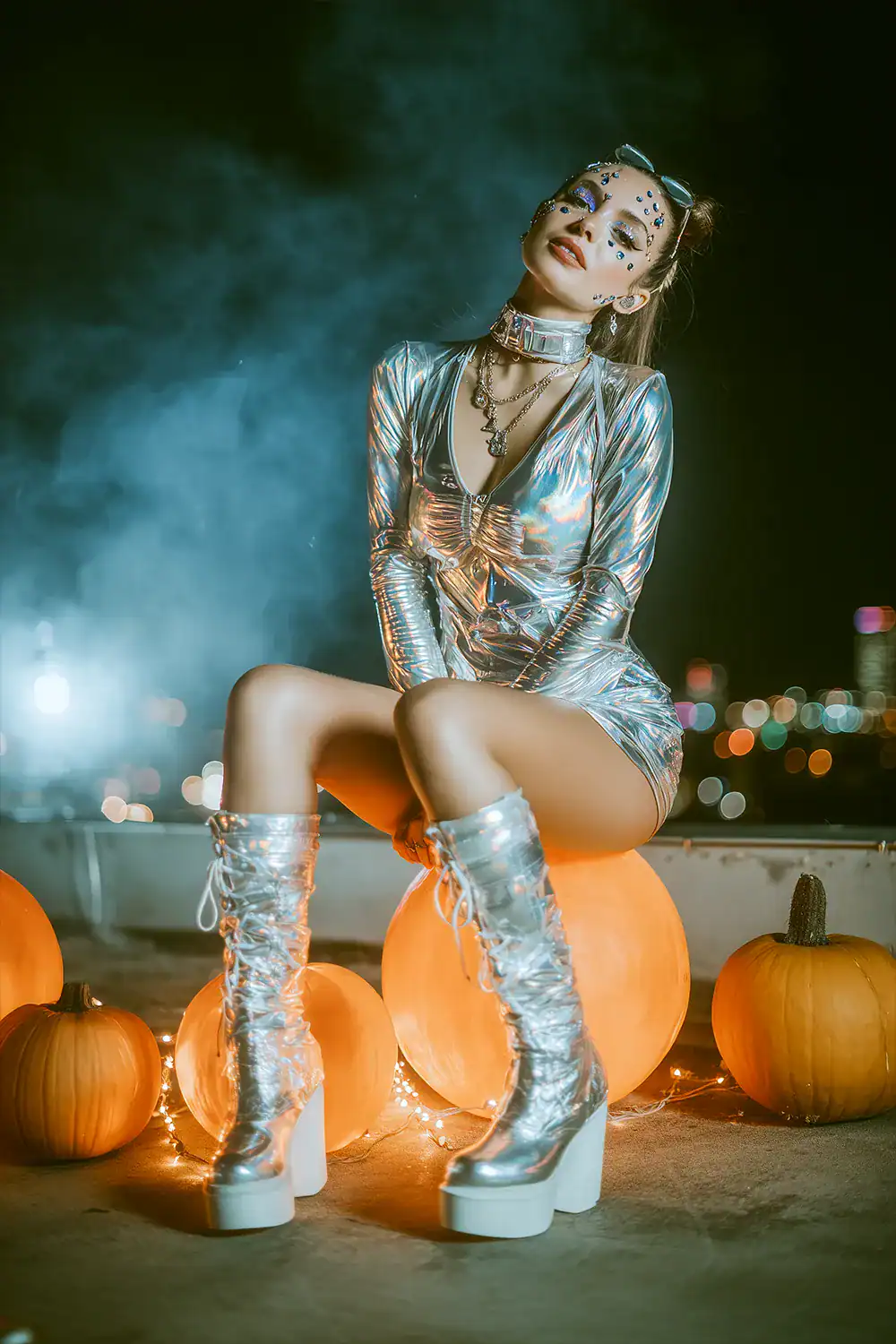 Frau in silbernem Metallic-Minidress mit kniehohen Plateaustiefeln und Face-Jewels, sitzt zwischen Kürbissen und Lichterkette auf einem Rooftop – Space-Babe Halloween-Kostüm für Frauen