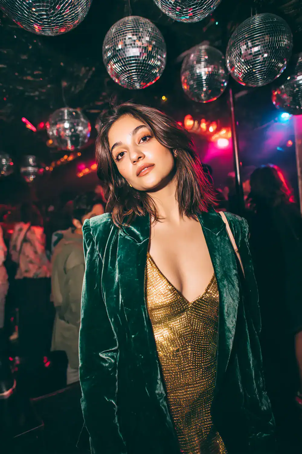 Frau im goldenen Pailletten-Kleid mit grünem Samtblazer auf einer glamourösen Silvesterparty mit Discokugeln